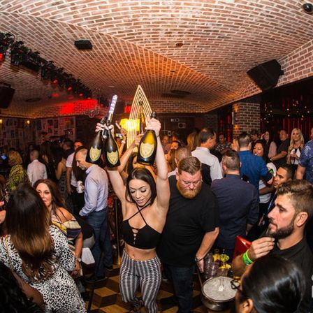 Lively nightclub lounge with arched brick ceiling and crowded dance floor as a server parades multiple champagne bottles overhead for bottle service
