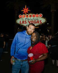 Couple grinning under the glowing "Welcome to Fabulous Las Vegas" sign at night — man in a blue hoodie holding a can, woman in a sparkly red dress with a red cup