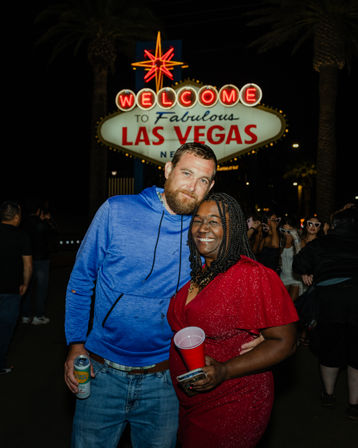 Couple grinning under the glowing "Welcome to Fabulous Las Vegas" sign at night — man in a blue hoodie holding a can, woman in a sparkly red dress with a red cup