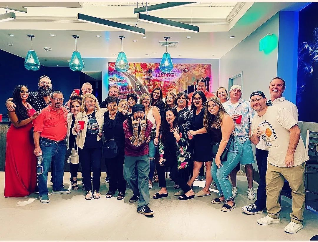 Large group of smiling adults posing at a cheerful indoor celebration in a modern lounge with turquoise pendant lights, a colorful mural and guests holding red cups.