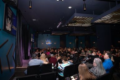Packed indoor comedy club audience seated at tables, watching a small stage with purple curtains and blue walls during a live comedy show under warm spotlights.
