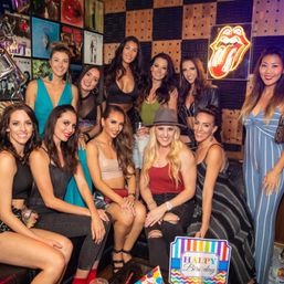 Group of women posing at a music-themed lounge for a birthday celebration, neon tongue sign and vinyl record wall art in the background.