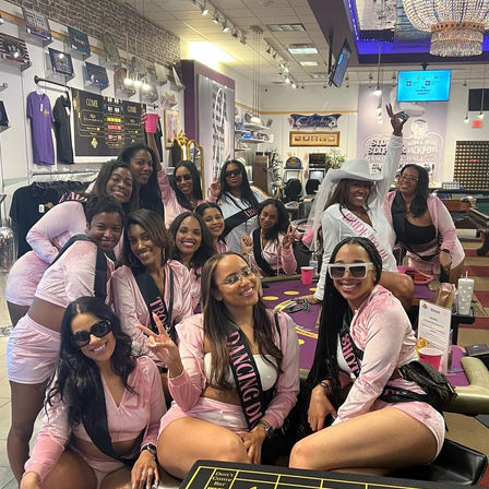 Smiling bachelorette party in matching pink outfits and sashes gathered around a casino table with slot machines and a sparkling chandelier overhead.