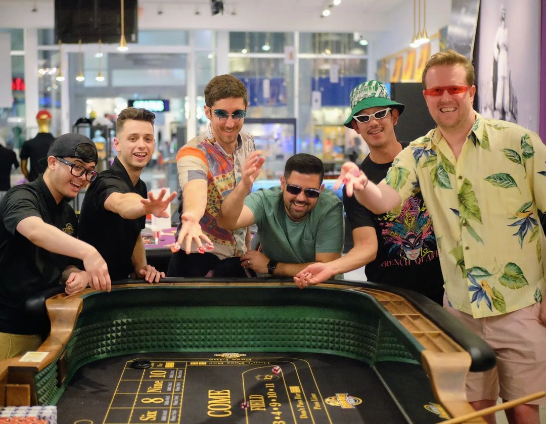 Six men in colorful shirts and sunglasses leaning over a craps table, laughing and reaching for dice in a bright indoor gaming room.