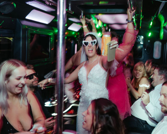 Bride in white wedding dress and heart-shaped sunglasses takes a selfie with a drink on a neon-lit party bus, surrounded by laughing friends in a lively bachelorette party nightlife scene