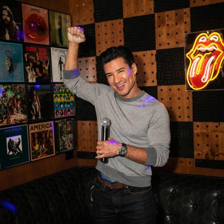 Smiling man celebrating with a microphone in a cozy karaoke bar booth, neon tongue sign and retro vinyl album covers on the wood-paneled wall