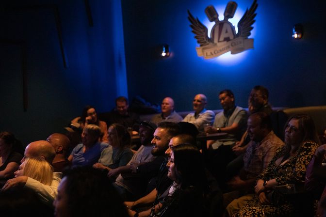 Laughing crowd in a dim, blue-lit comedy club watching a stand-up show, lively nightlife audience with illuminated microphone logo on the wall