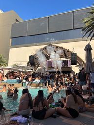 Vibrant daytime dayclub pool party at a sunny resort, packed swimmers and loungers, DJ stage with smoke effects, palm trees and blue sky