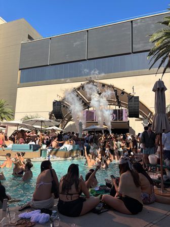 Vibrant daytime dayclub pool party at a sunny resort, packed swimmers and loungers, DJ stage with smoke effects, palm trees and blue sky