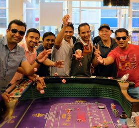 Group of men cheering and tossing dice at a lively casino craps table with purple felt, poker chips and drinks