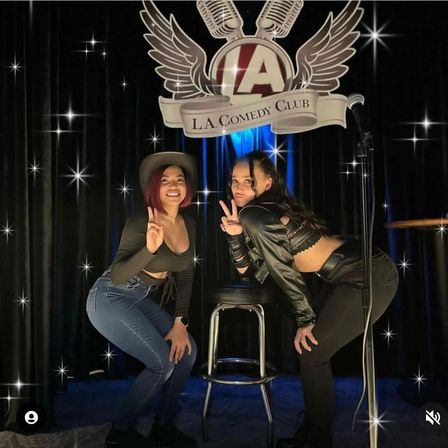 Two women flashing peace signs and posing on a small comedy club stage with a microphone stand, bar stool and starry curtain backdrop — casual night out at a live comedy venue.