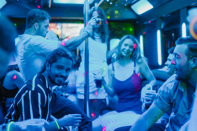Friends partying inside a neon-lit party bus, laughing and holding drinks around a dance pole—energetic nightlife scene