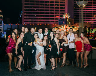 Group of friends partying on a neon-lit Las Vegas casino strip at night — bachelorette-style celebration with cocktails, sequined and cocktail dresses, and a woman in a white dress and sneakers.