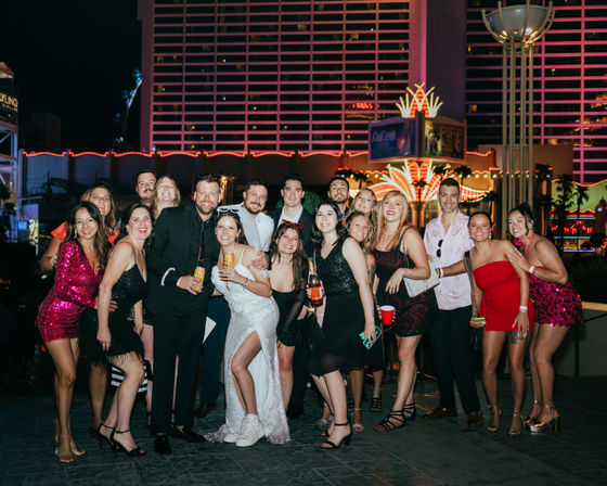 Group of friends partying on a neon-lit Las Vegas casino strip at night — bachelorette-style celebration with cocktails, sequined and cocktail dresses, and a woman in a white dress and sneakers.