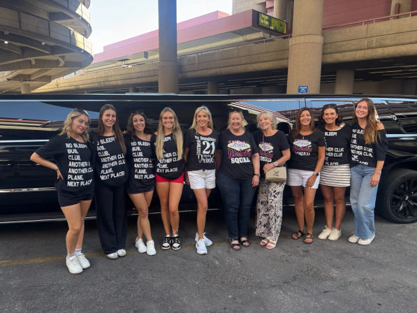 Group of ten women in matching celebratory tees and casual summer outfits posing in front of a black stretch limousine outside an urban parking ramp, lively Las Vegas birthday getaway vibe.