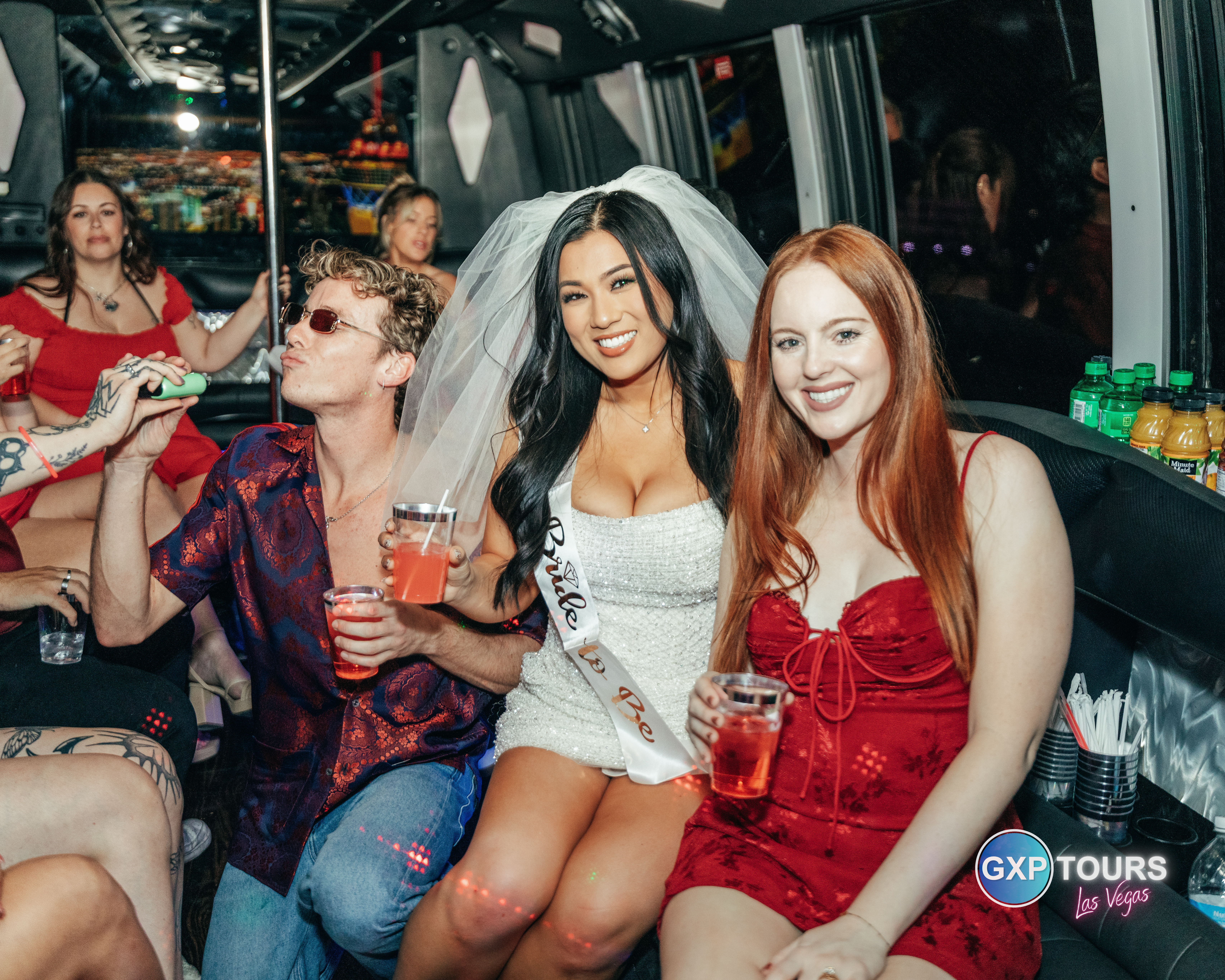 Bachelorette party on a Las Vegas party bus: bride-to-be in a white dress, veil and sash smiling between friends in red and patterned outfits, all holding drinks and enjoying the night ride.