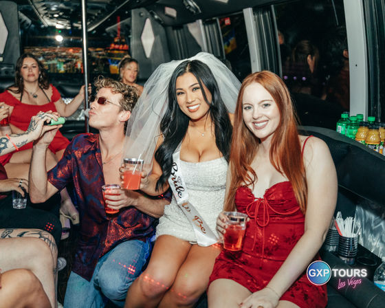 Bachelorette party on a Las Vegas party bus: bride-to-be in a white dress, veil and sash smiling between friends in red and patterned outfits, all holding drinks and enjoying the night ride.