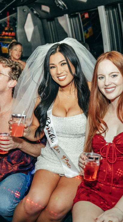 Bachelorette party on a Las Vegas party bus: bride-to-be in a white dress, veil and sash smiling between friends in red and patterned outfits, all holding drinks and enjoying the night ride.