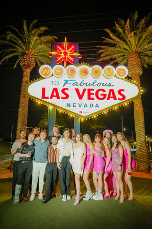 Group of friends posing at night beneath the illuminated "Welcome to Fabulous Las Vegas Nevada" sign with palm trees, many wearing bright pink party outfits.