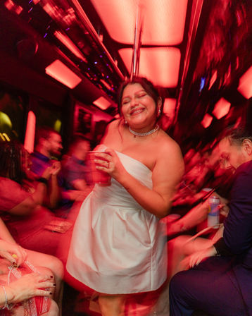 Smiling woman in a white cocktail dress holding a red cup, dancing inside a red-lit party bus with blurred friends around in a lively celebration.
