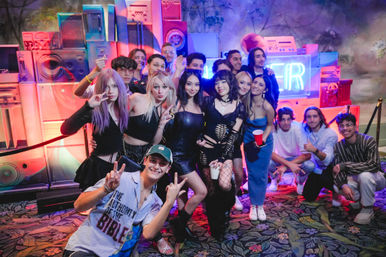 Neon-lit indoor party: a diverse group of young adults posing and smiling in front of a colorful speaker-wall backdrop, holding drinks and making peace signs in a lively nightclub-style lounge.