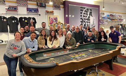 Twenty adults smiling and posing around a casino-style craps table inside a game and entertainment shop, with merchandise shelves, arcade machines, and gold celebration balloons in the background.