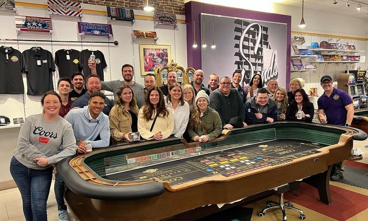 Twenty adults smiling and posing around a casino-style craps table inside a game and entertainment shop, with merchandise shelves, arcade machines, and gold celebration balloons in the background.