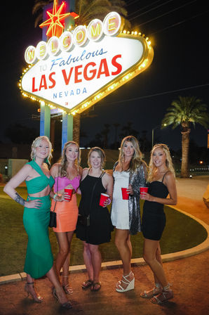Five friends posing at night beneath the illuminated Welcome to Fabulous Las Vegas Nevada sign, holding red cups with palm trees in the background
