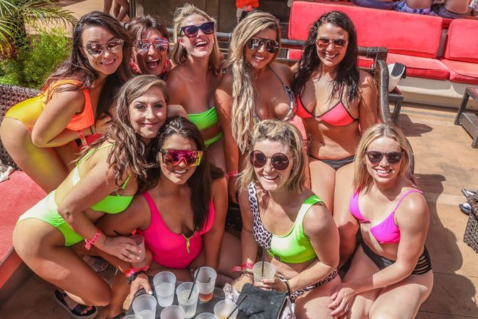 Smiling group of women in bright swimsuits and sunglasses posing together on a sunny pool-deck lounge with drinks and red cushioned seating