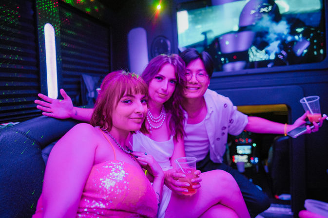 Three friends smiling and holding drinks in a neon-lit party bus lounge, purple and blue lights and laser dots creating a lively nightlife vibe.
