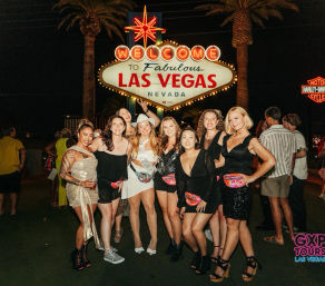 Group of women posing and celebrating at night beneath the illuminated “Welcome to Fabulous Las Vegas Nevada” sign, wearing party outfits and colorful fanny packs with palm trees and neon lights in the background.