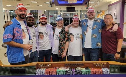 Seven friends in matching bachelor‑party T‑shirts and Las Vegas visors posing at a casino poker table stacked with colorful chips