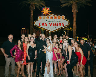 Bride and groom with a large group of friends in festive outfits posing at night beneath the iconic neon Las Vegas sign, palm trees and city lights in the background.