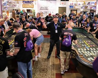 Crowded casino gaming event around large craps tables with players and dealers, a man in the center raising his arms while others place bets and cheer.
