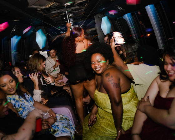 Party bus interior at night with colorful LED lights, a woman in a lime-green dress smiling at the camera, another dancing on a center pole, and a lively group of people seated with drinks and phones celebrating.