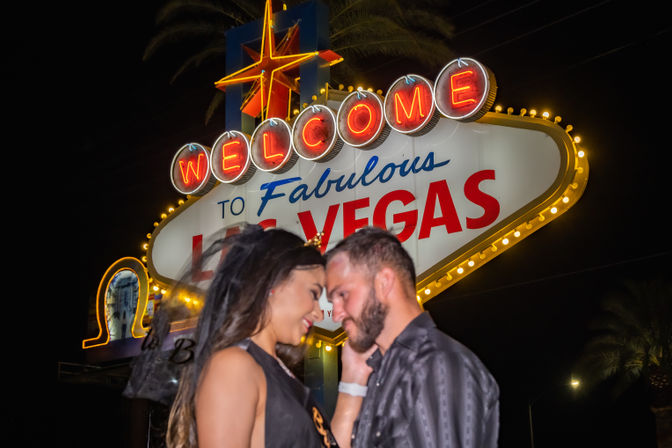 Romantic couple forehead-to-forehead at night beneath the illuminated "Welcome to Fabulous Las Vegas" neon sign with palm trees and glowing marquee lights.