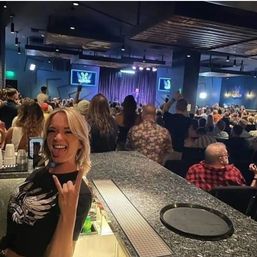 Smiling blonde woman at a bar flashing a rock hand sign in a crowded indoor comedy club, audience watching a lit stage with curtains and screens — lively live-show nightlife scene.