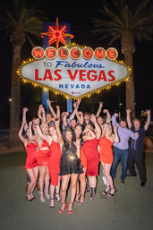 Group of friends posing at night under the illuminated "Welcome to Fabulous Las Vegas Nevada" sign, wearing red and black party outfits with palm trees and neon lights in the background.