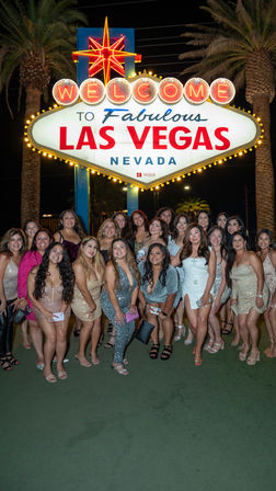 Large group of women in sparkly party outfits posing at night in front of the illuminated Welcome to Fabulous Las Vegas Nevada sign with palm trees and neon lights