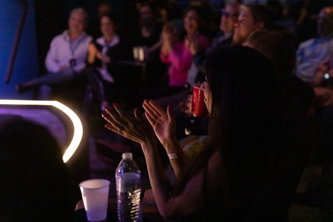 Crowd cheering at a live comedy club show — front-row patron clapping beside a water bottle and cups near an illuminated stage edge.