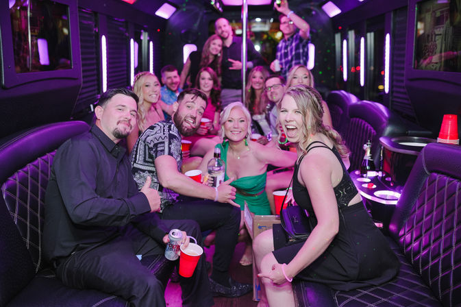 Lively group of friends celebrating inside a purple-lit party bus, laughing and posing around a dance pole with red cups and bottles for a fun nightlife party vibe.
