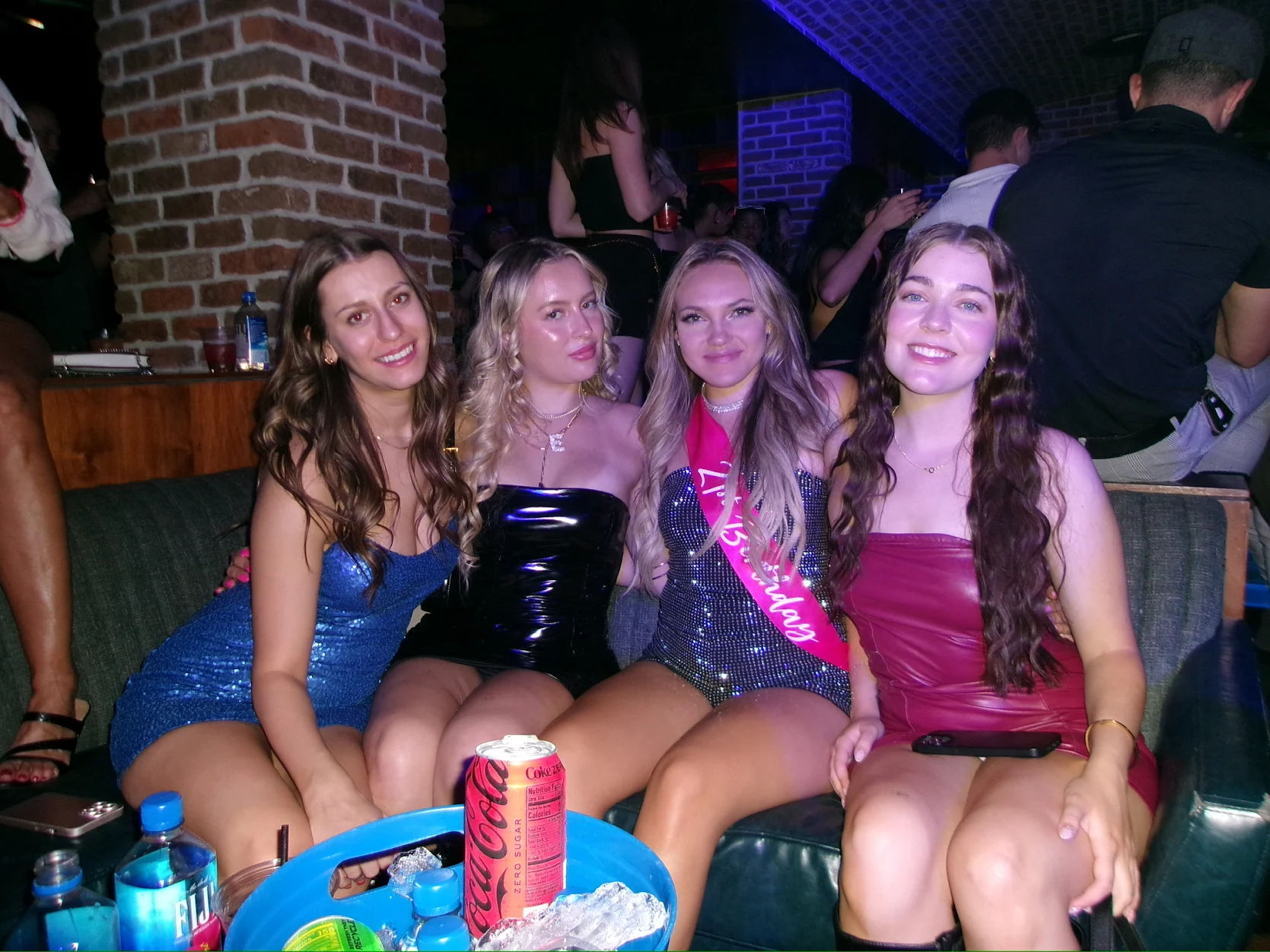Four women smiling on a sofa in a brick-walled nightclub lounge, wearing sparkly party dresses with a pink birthday sash on one; bottles and a Coca-Cola can on the table in front.