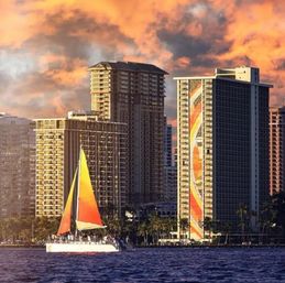 Bright orange-sailed catamaran cruising on blue water in front of a palm-lined waterfront and tall hotel skyscrapers under dramatic orange sunset clouds.