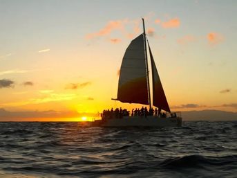 Silhouette of a catamaran sunset cruise with passengers on deck sailing across rippling ocean waves under a golden-orange sky