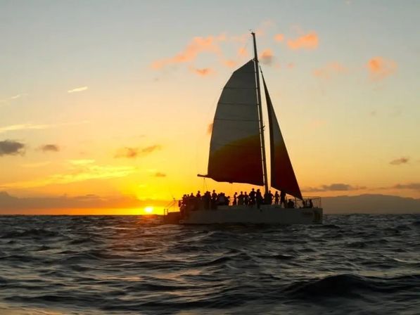 Silhouette of a catamaran sunset cruise with passengers on deck sailing across rippling ocean waves under a golden-orange sky