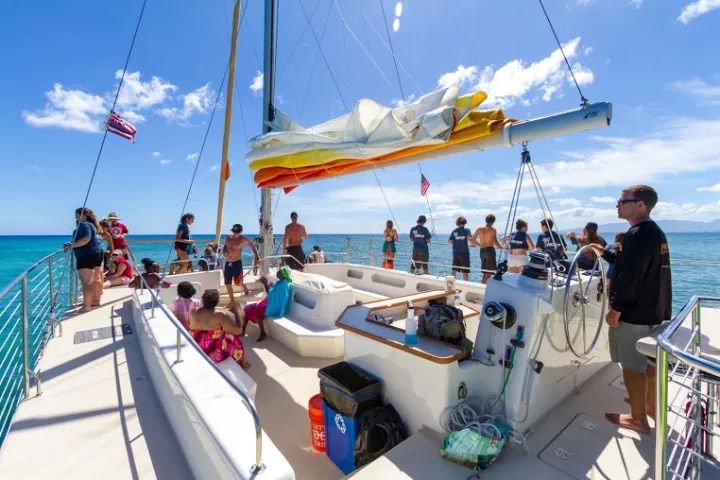 Group of people on the deck of a white catamaran sailboat in turquoise tropical waters on a sunny day, folded colorful sail above and crew at the helm.