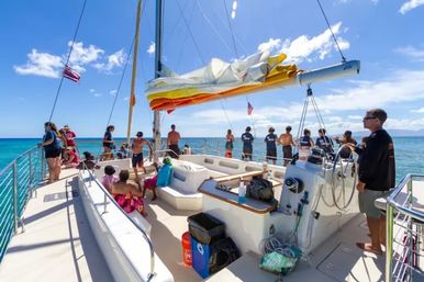 Group of people on the deck of a white catamaran sailboat in turquoise tropical waters on a sunny day, folded colorful sail above and crew at the helm.