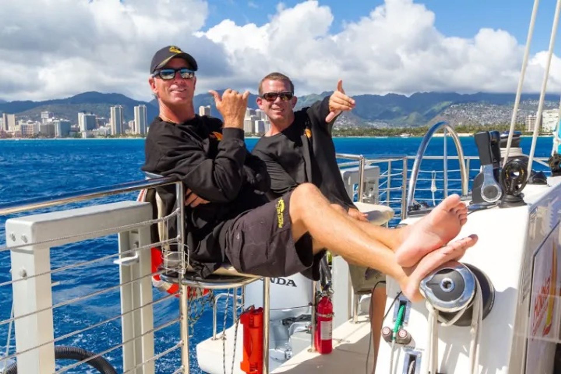 Two relaxed crew members in sunglasses and black shirts flashing shaka signs and lounging barefoot on a sunny sailboat deck with deep-blue ocean and a tropical coastal skyline and mountains in the background.