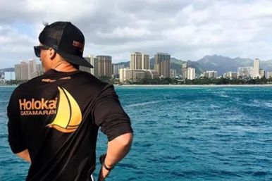 Crew member in a black cap and shirt with a yellow sail logo gazes across turquoise ocean toward a sunlit coastal city skyline from a boat.
