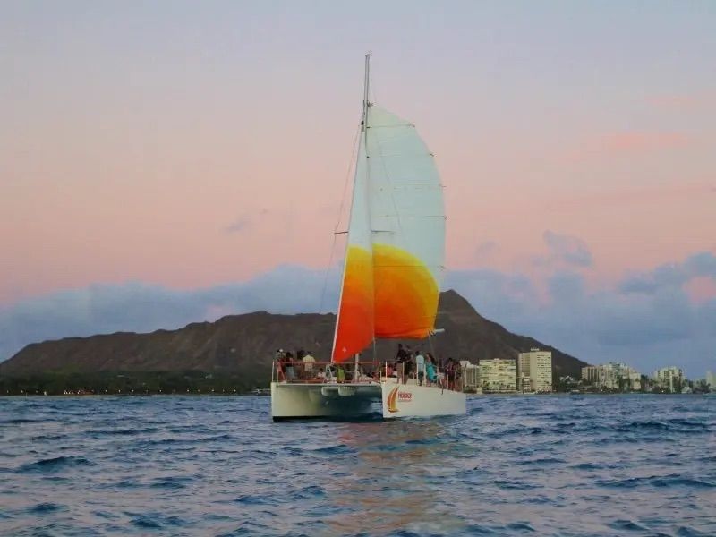 Sunset catamaran with a bright orange-yellow sail cruising off Waikiki with Diamond Head crater and Honolulu skyline in the background